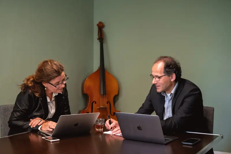 Ute Lysk und Dr. Georg Barzel im Gespräch an einem Besprechungstisch mit Laptops, im Hintergrund ein Kontrabass